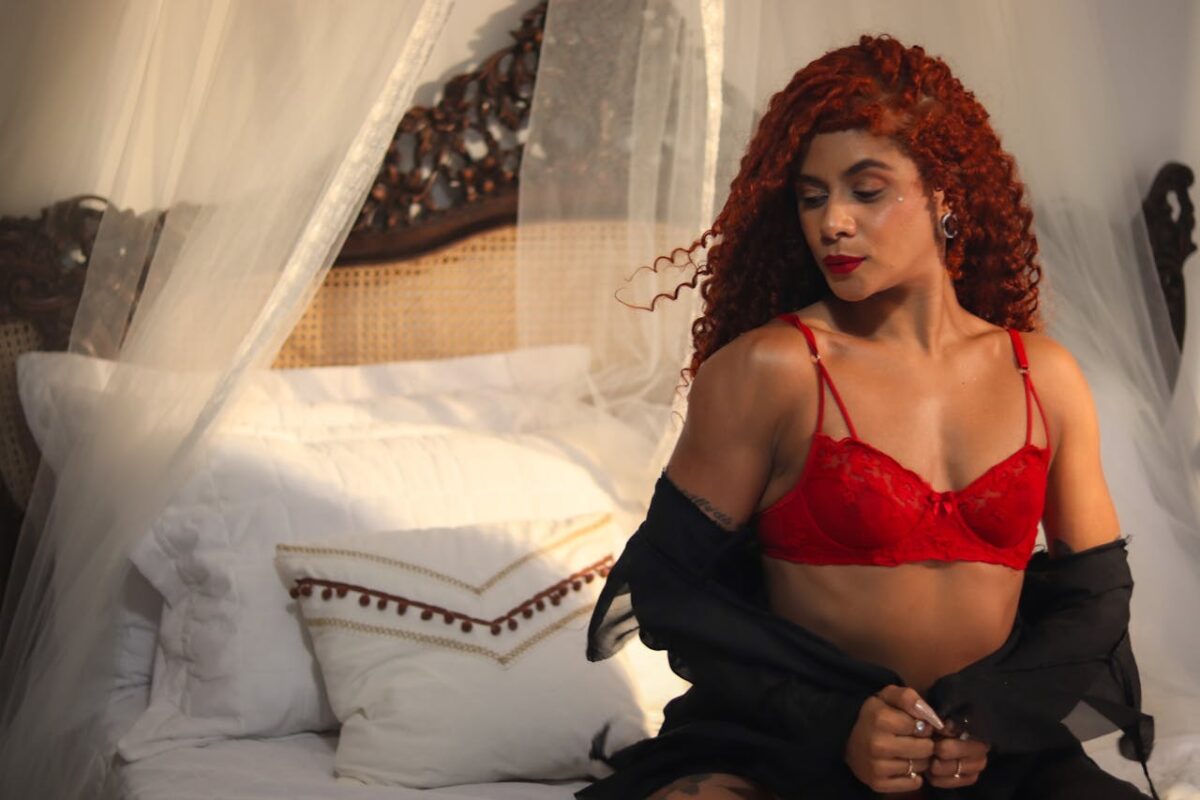 A woman in red lingerie poses elegantly on a vintage carved bed with white bedding.