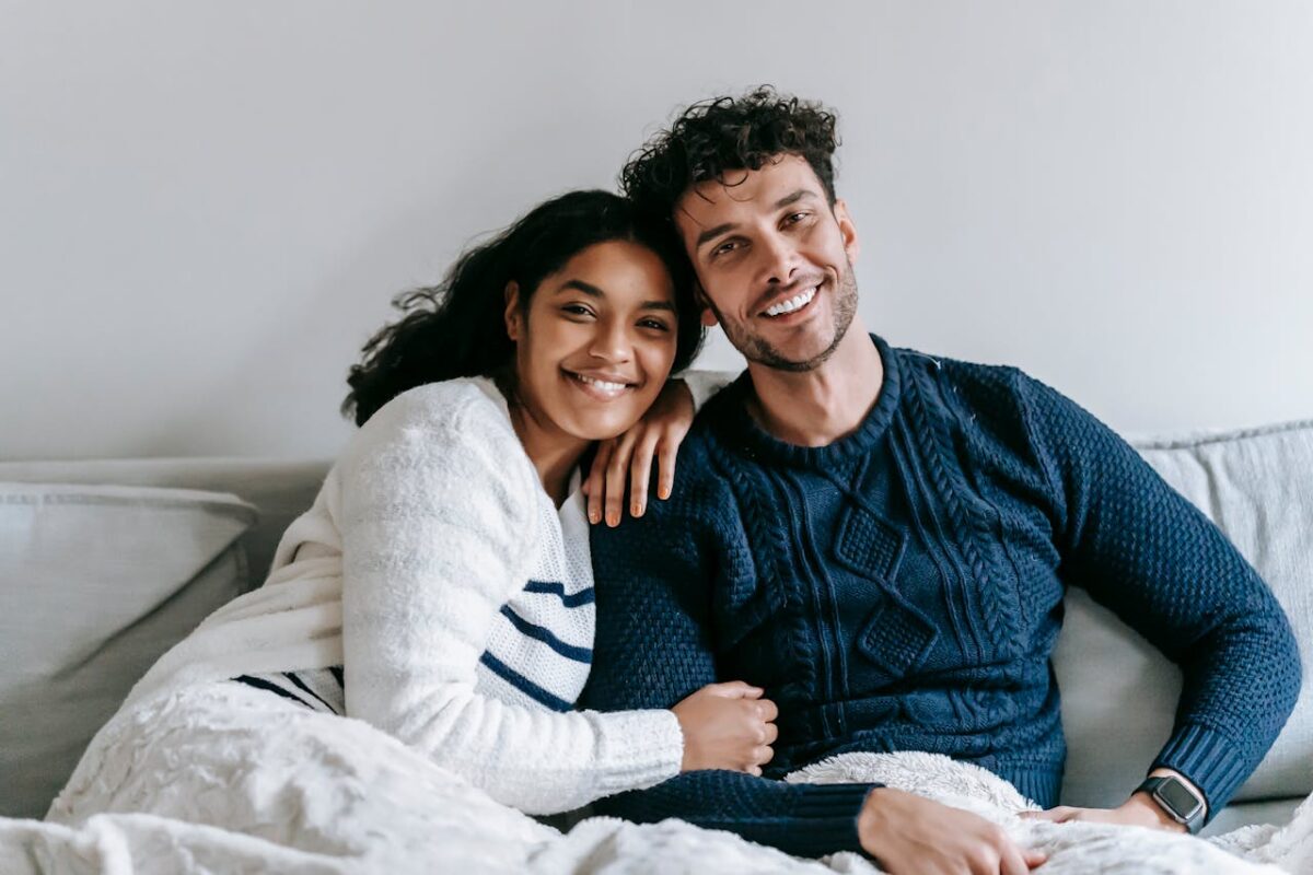 Services Happy ethnic couple in casual clothes sitting on soft sofa in light room and looking at camera in daytime